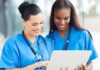 Two nurses in blue scrubs smiling while looking at a tablet
