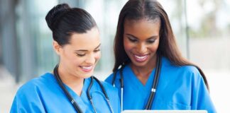 Two nurses in blue scrubs smiling while looking at a tablet