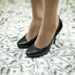 A pair of black high heels standing on a floor covered with dollar bills